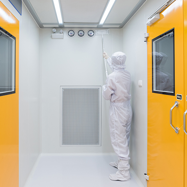 Worker in protective coveralls cleaning the upper wall of a cleanroom with a long-handled mop.