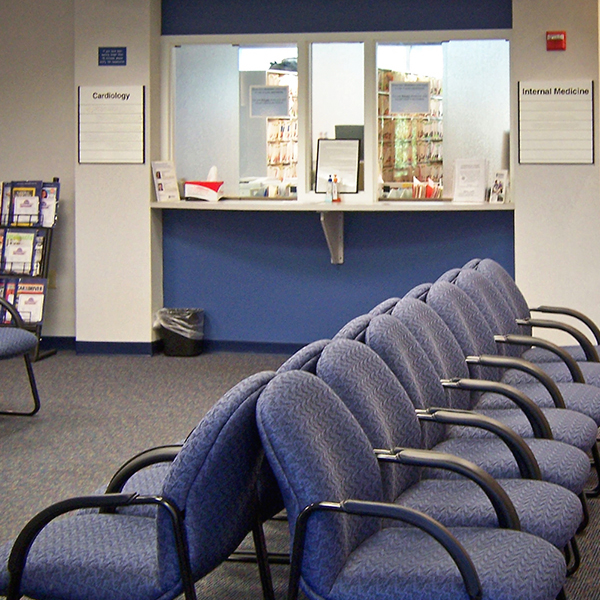 Medical office waiting room with rows of blue upholstered chairs and a reception window.