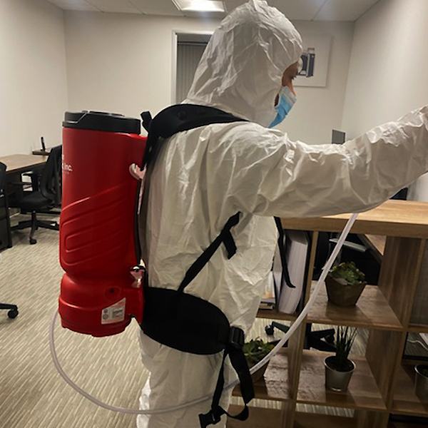 Worker wearing protective coveralls, a face mask, and a red backpack sprayer in an office.
